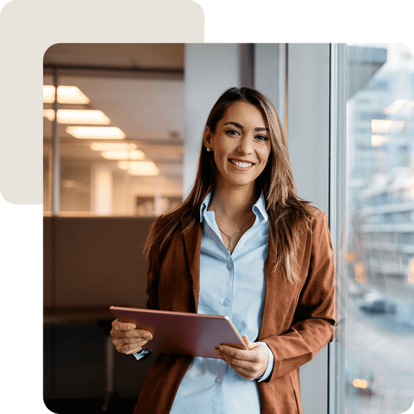 Geschäftlich gekleidete Frau mit Tablet in der Hand lehnt lächelnd an einer Fensterscheibe.