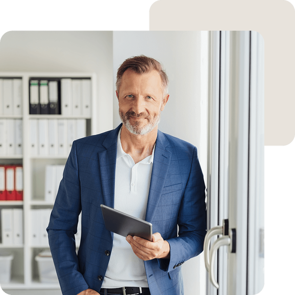 Mann im Sakko steht lächelnd in einem Büro vor einem Regal mit Ordnern und hält ein Tablet in der Hand.