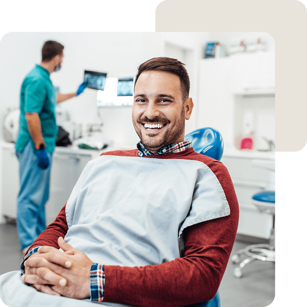 Mann sitzt lächelnd auf dem Zahnarztstuhl während der Zahnarzt Röntgenaufnahmen betrachtet